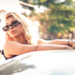 Stylish blonde woman wearing sunglasses while relaxing outside a car on a sunny day.